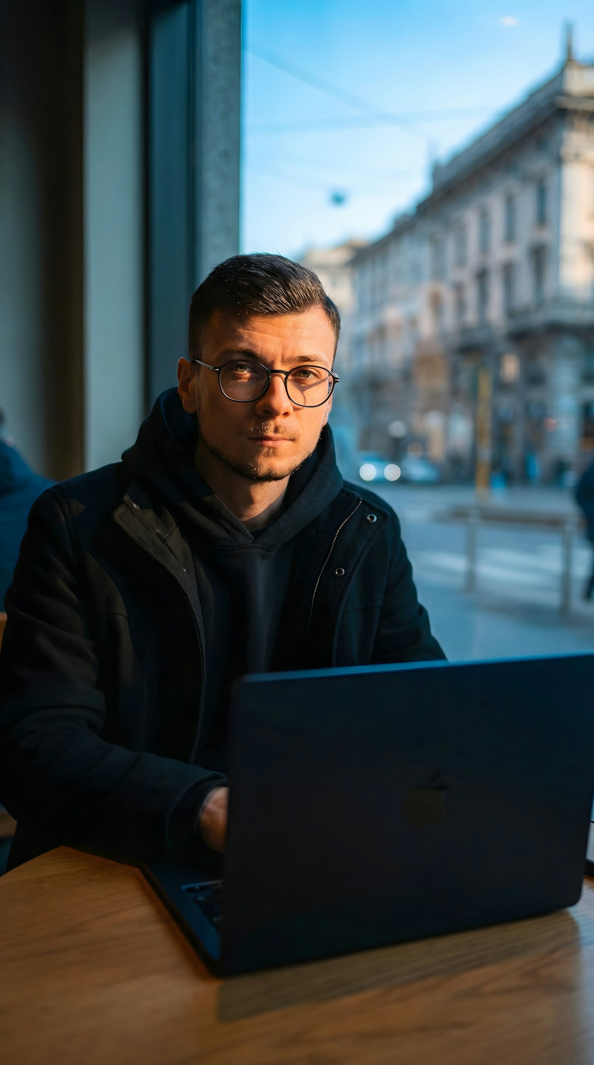 A professional with glasses at a café window, MacBook open, golden light — focused, calm, in control